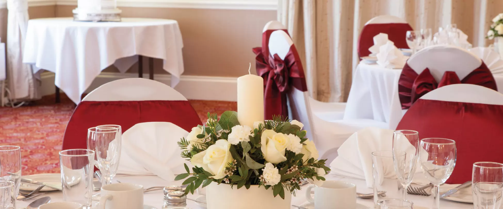 A circular table set up for a wedding breakfast with white table cloths, red ribbons and bouquets of white flowers..